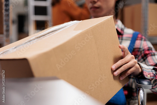 Warehouse worker using wheelchair while holding cardboard box in hands. Storehouse order handler preparing product for dispatching, taking packed parcel ready for shipment close up photo