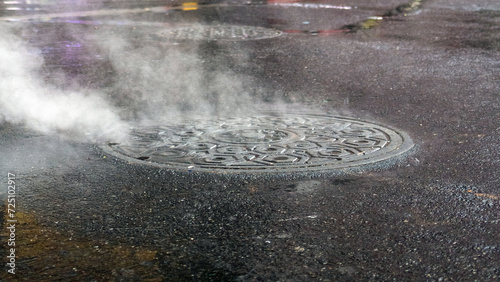 A manhole cover lets off steam and white smoke on a wet asphalt road after rain has finished during hot and humid weather