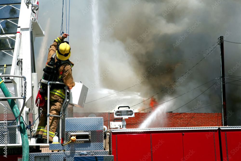 A fireman standing on a hook and ladder fire engine and giving directions to other firefighters where to spray the water while battling a blazing structure fire.
