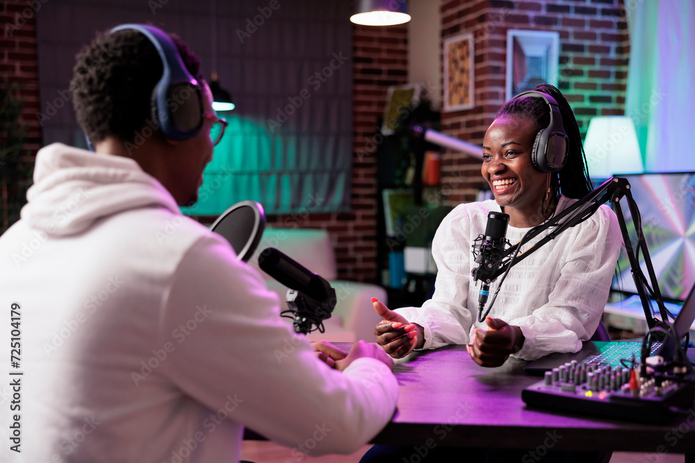 Online show host interviewing african american woman making him laugh during live broadcast. Talking show interviewer using professional microphone and decor to record comedy podcast