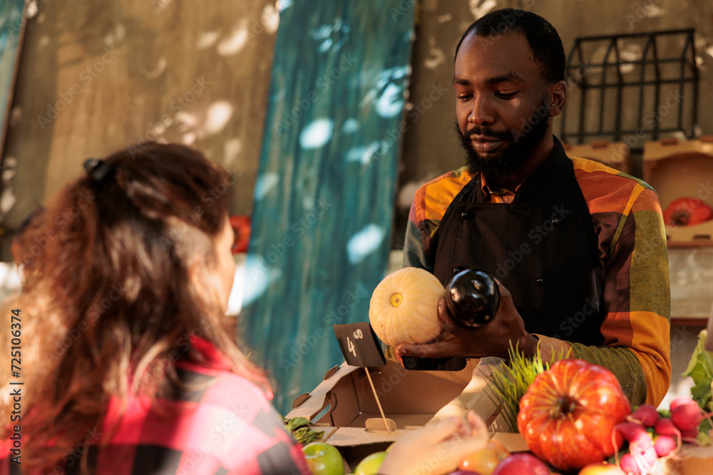 Young local seller showing organic healthy products from farmers market stall, locally grown food. Male farmer talking to customer about various seasonal fresh veggie produce.