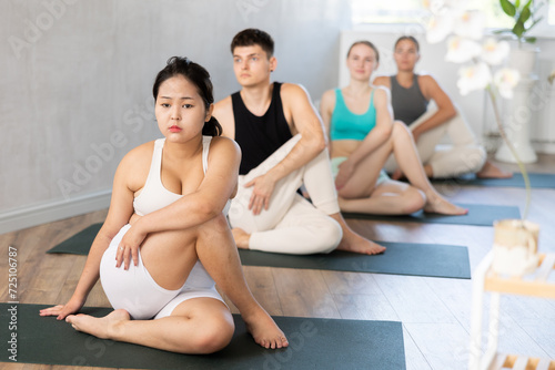 Focused young Asian teen girl sits on karemat, looks towards teacher and performs Lord of Fishes asana by Ardha Matsyendrasana. Young people do hatha yoga in background