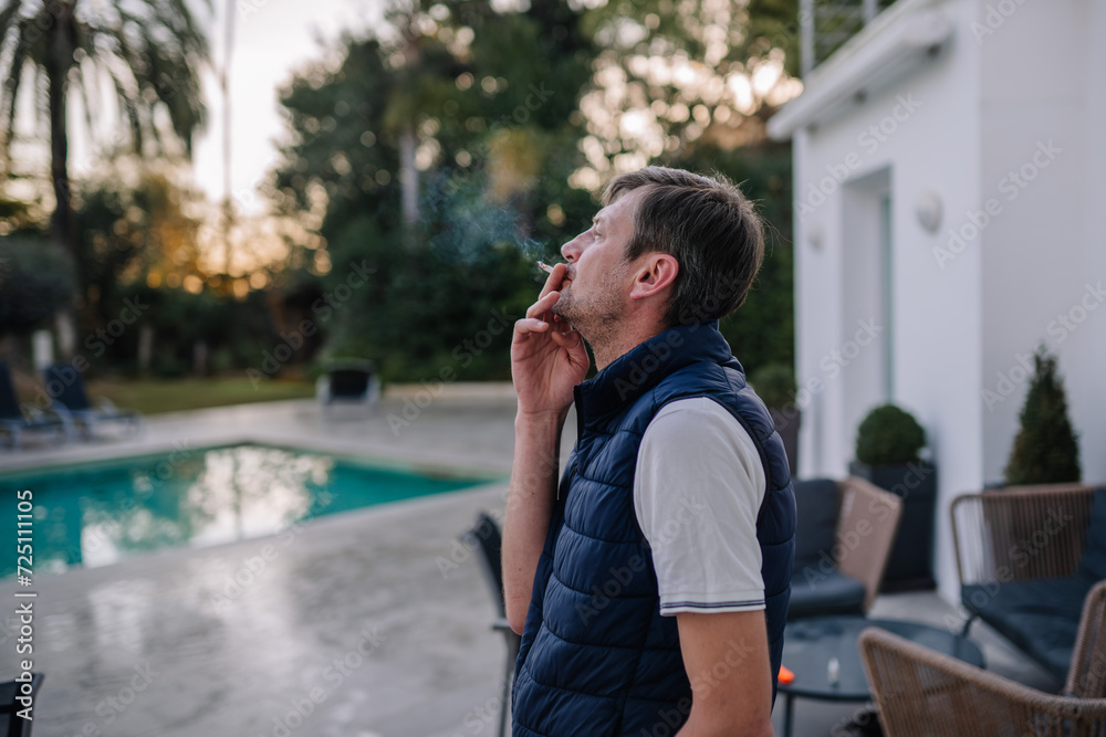 Man smoking outdoors, blurred pool background, dusk, trees, patio chairs, vest, casual.