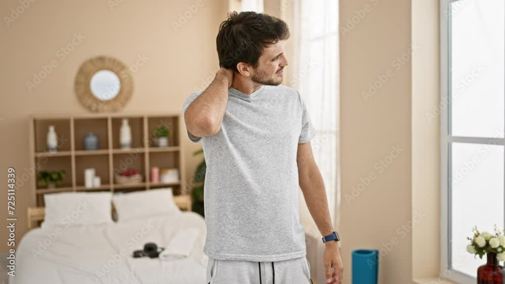 A young man massages his neck feeling discomfort in a well-lit bedroom with simple decor.