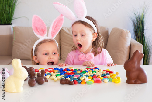 Small children, kids sisters with rabbit ears hunt for chocolate eggs eat them and laugh celebrate on the table in home.