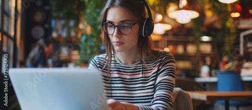 Motivated young woman in striped shirt observing computer screen and using tablet. Innovative girl in headphones and glasses working in office post coffee break.