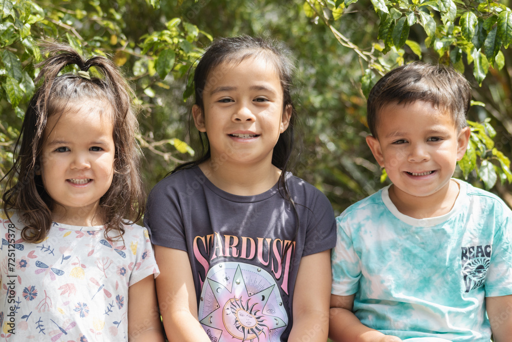 a close up image of three maori children sitting for their family portrait outdoors in a natural setting. - obrazy, fototapety, plakaty 