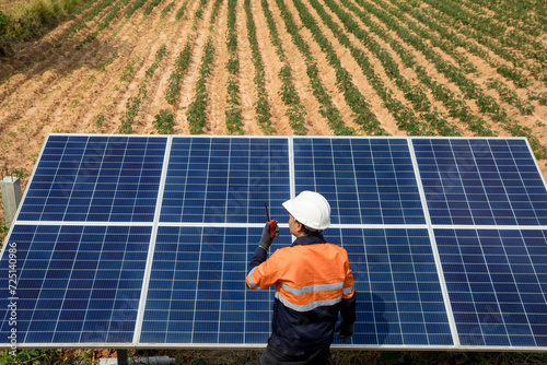 A young male technician in work clothes uses a walkie-talkie while inspecting a solar panel system in the field.