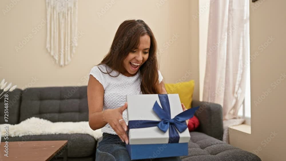 Radiating joy, a young hispanic woman sitting comfortably on her sofa at home, confidently unpacking a surprise birthday gift, her beautiful smile expressing pure happiness.