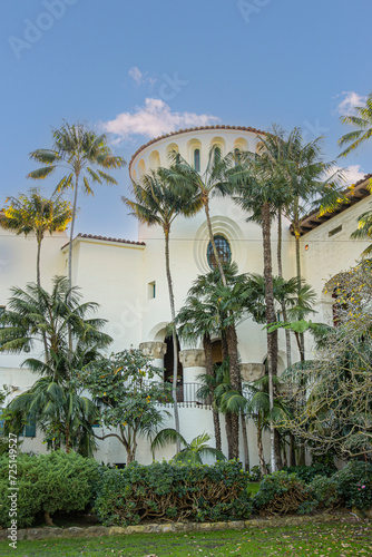 Santa Barbara, CA, USA - November 30, 2023: Santa Barbara County Courthouse garden side view on white stone circular south tower against blue sky. Tall green palm trees and plants