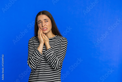 Dental problems. Young woman touching cheek, closing eyes with expression of terrible suffer from painful toothache sensitive teeth cavities. Pretty girl isolated on blue studio background. Copy-space