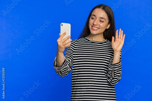 Happy Caucasian woman blogger taking portrait selfie on smartphone, streaming vlog, communicating record video call online with social media subscribers. Girl isolated on blue background. Copy-space © Andrii Iemelianenko