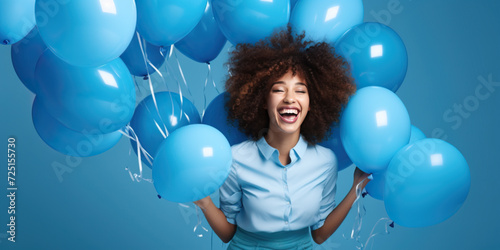 Cheerful curly haired woman dressed in blue clothes, laughing around inflated helium balloons over blue background