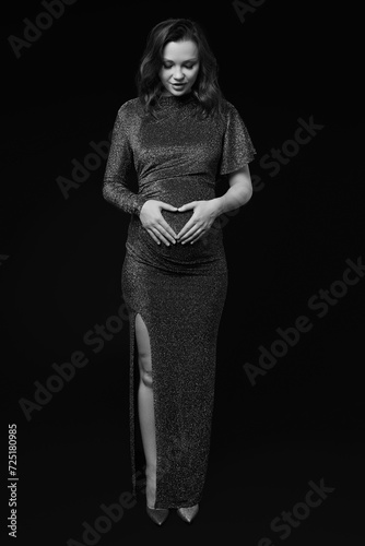 Black and white portrait of pregnant female in sequin dress with hands near pregnant belly with heart sign.