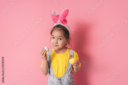 Little girl with bunny ears on her head and Easter eggs in her hands against a pink background.