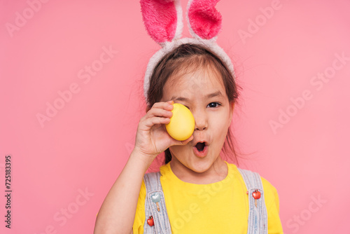 Little girl wearing bunny ears on her head holding yellow Easter egg in her hand against pink background.
