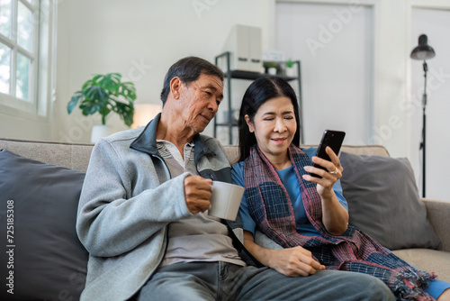 Retired elderly couple sits on couch drink tea and using mobile together and relax in their home. Senior Activity Concept photo