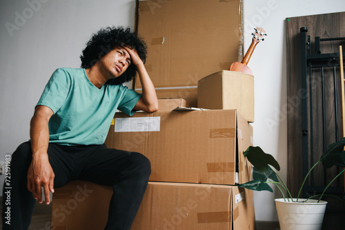 Asian man touching his head and sitting near stacked of carton boxes with sad expression photo
