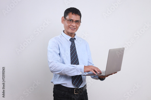 Adult Asian businessman wear glasses and formal clothes holding laptop while looking at camera smiling