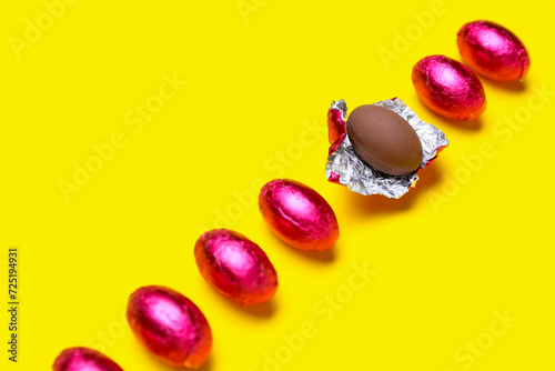 Unpacked chocolate Easter egg among wrapped ones on yellow background, closeup