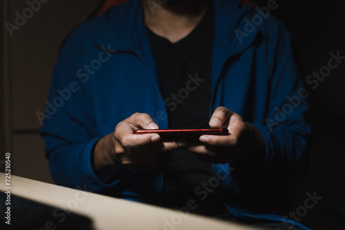 A man in a blue shirt uses a smartphone or tablet in a dark room. Holding hands. Black background. Home office. For work. For social media or searching for information