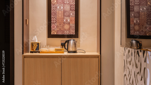 A place for making tea and coffee. There is an electric kettle, cups, glasses, napkins on the table in the corner of the room. An ornament on a beige wall.