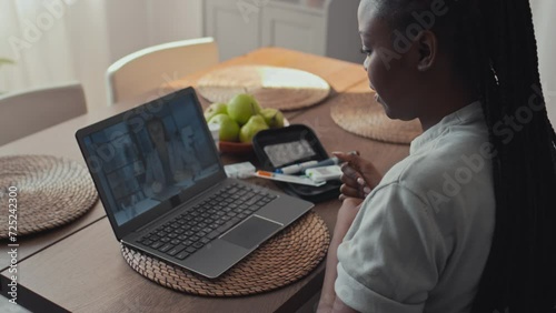 Medium shot of African American woman with diabetes sitting in front of laptop, having telemedicine appointment with GP on video call, reporting on her condition, showing glucose monitor device on arm photo