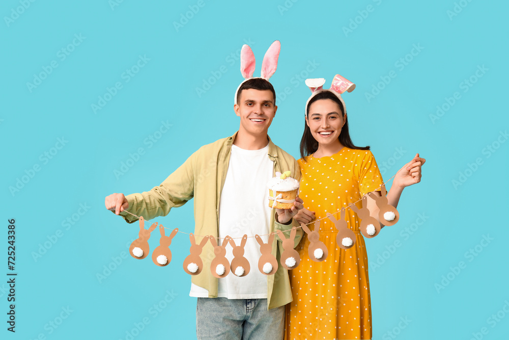 Happy young couple with Easter bunny ears, cake and garland on blue background
