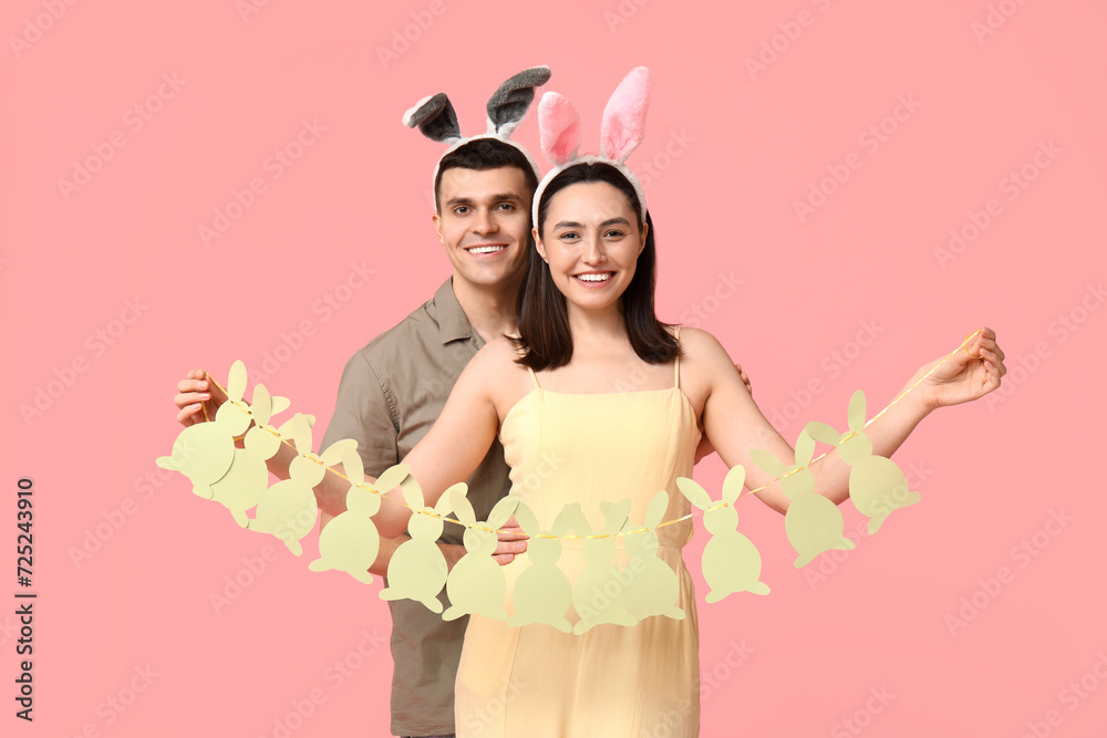 Happy young couple with Easter garland and bunny ears on pink background Happy young couple with Easter garland and bunny ears on pink background