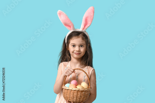Cute little girl in bunny ears with basket of Easter eggs on blue background