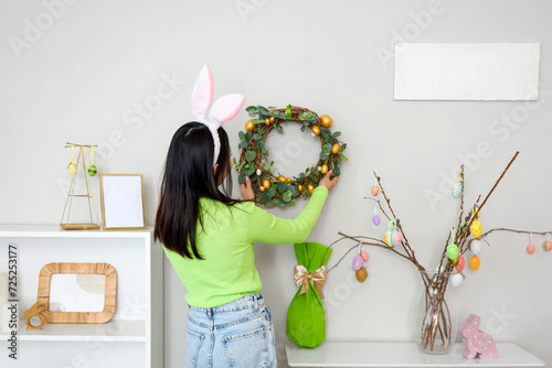 Young Asian woman in bunny ears hanging Easter wreath on wall at home