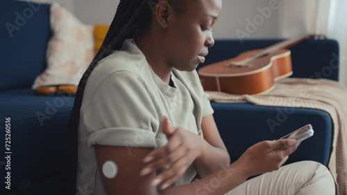 Medium close-up side shot of young African American woman with diabetes sitting on floor by couch in living room, touching glucose monitoring device on arm, then saving data on smartphone app photo