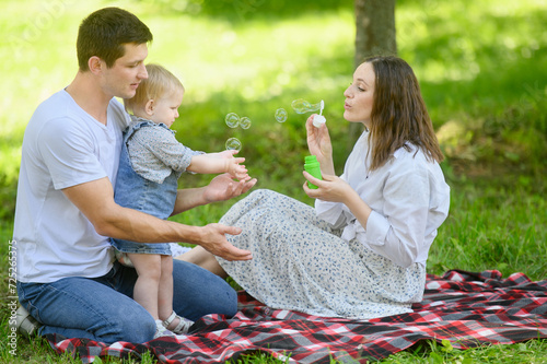 Cute young family sits on a blanket in park, mother blows soap bubbles, father and child catch them, play, have fun togetheron a warm sunny weekend. © GRON777