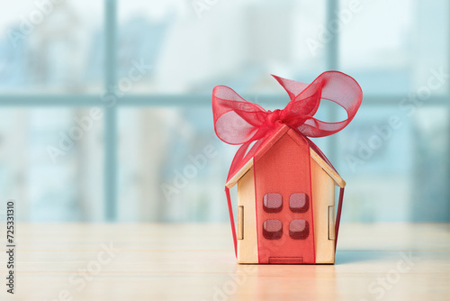 Small house gift with red bow over defocused cityscape with copy space. Concept of real estate gift or purchasing