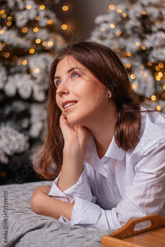 A young woman in a white shirt and blue jeans lies on a bed against a background of Christmas trees and bokeh of garlands.