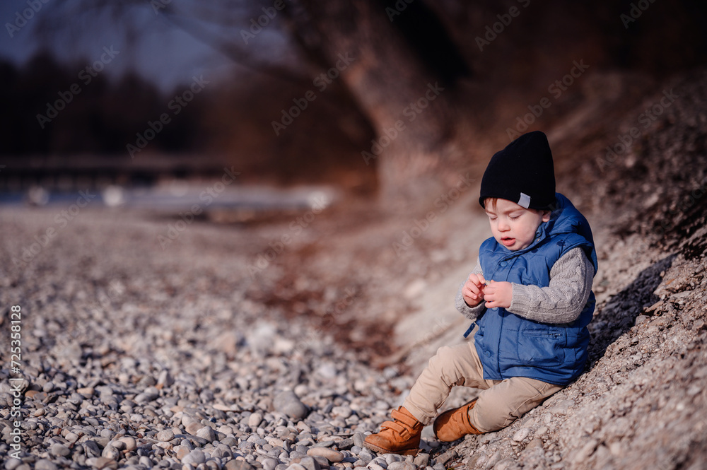 Curious toddler sitting on pebbles by the lake, exploring with a focused expression Curious toddler sitting on pebbles by the lake, exploring with a focused expression