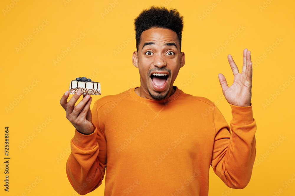 Young sad mad man wear orange sweatshirt casual clothes hold in hand piece of cake dessert spread hand isolated on plain yellow background. Proper nutrition healthy fast food unhealthy choice concept.