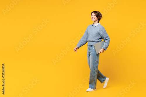 Full body side view young IT woman she wears grey knitted sweater shirt casual clothes hold use work on laptop pc computer walk isolated on plain yellow background studio portrait. Lifestyle concept.