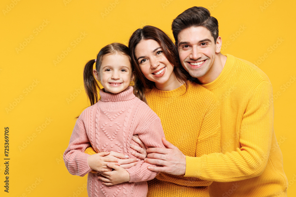 Close up young fun happy parents mom dad with child girl 7-8 years old wearing pink knitted sweater casual clothes hug cuddle look camera isolated on plain yellow wall background. Family day concept.