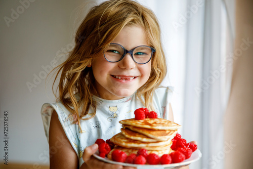 Little happy preschool girl with a large stack of pancakes and raspberries for breakfast. Positive child eating healthy homemade food in the morning.