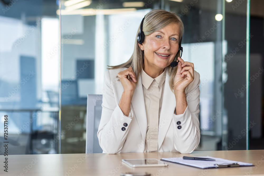Happy elderly female in light formal clothes in wireless headset setting microphone by table in office space. Blue eyed woman working as customer support representative and providing service. Happy elderly female in light formal clothes in wireless headset setting microphone by table in office space. Blue eyed woman working as customer support representative and providing service.