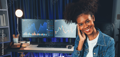 Profile of young African American businesswoman smiling on happy face wearing jeans shirt, sitting on chair against stock exchange market screen background. Concept of investment blogger. Tastemaker. photo
