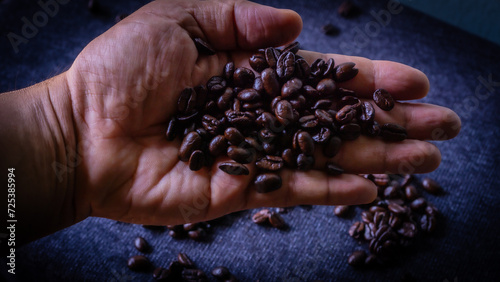 Black, dark brown coffee beans rest in the palm of your hand. With some coffee beans placed on a black cotton cloth in the background.