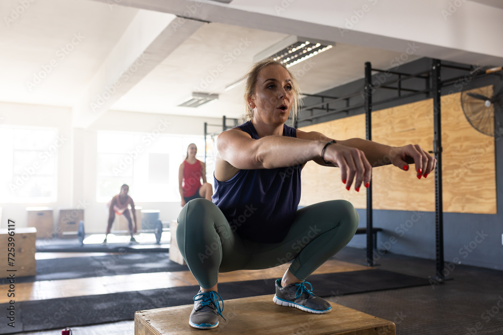 Full length of dedicated caucasian woman practicing squats on wooden plyo box in health club Full length of dedicated caucasian woman practicing squats on wooden plyo box in health club