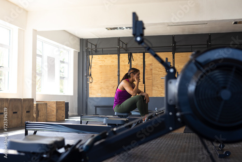 Side view of exhausted biracial young woman sitting on rowing machine in health club, copy space photo