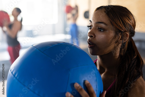 Confident biracial young woman exercising with blue fitness ball in heath club, copy space photo