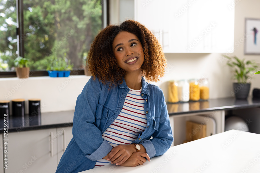 Happy biracial woman with curly hair leaning on counter smiling in kitchen Happy biracial woman with curly hair leaning on counter smiling in kitchen