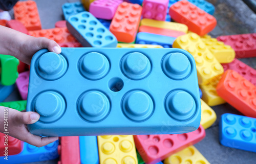Kind hält einen großen blauen Bauklotz über einen Haufen vieler weiterer bunter XXL-Spielbausteine aus Kunststoff draußen in einem Spielbereich