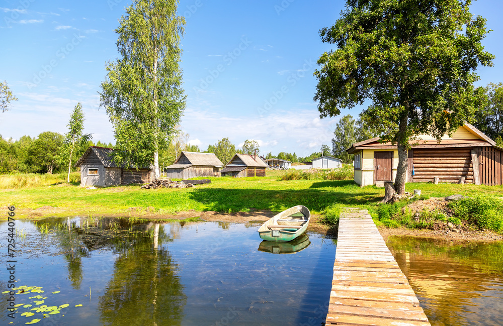 Fototapeta premium Old fishing plastic boat moored at a pier on the lake and traditional russian wooden baths on the shore in the summer