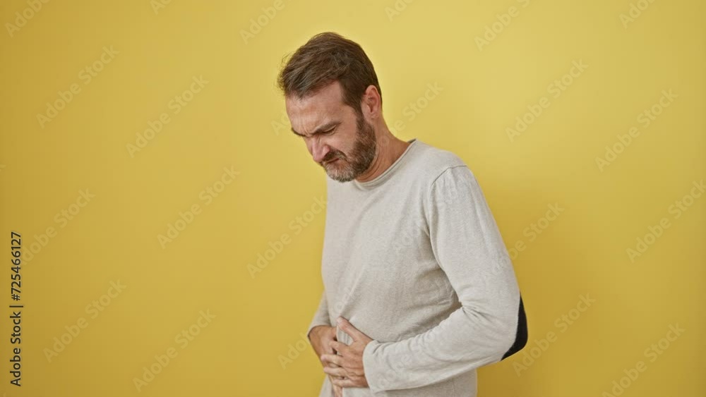 Handsome middle-aged hispanic man in pain, standing with hand on stomach, nauseous from a painful disease; a sick expression against isolated yellow background